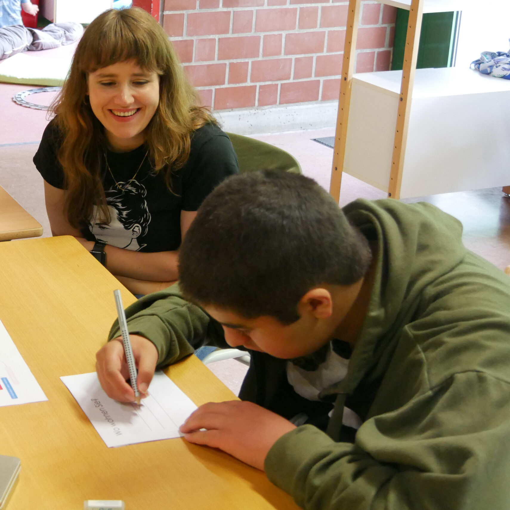 Ricarda Lang, eine Frau mit langen braunen Haaren, lächelt, während sie einem Jungen in einem grünen Kapuzenpulli dabei zusieht, wie er an einem Tisch in einem Klassenzimmer auf ein Blatt Papier schreibt. Ricarda Lang, eine Frau mit langen braunen Haaren, lächelt, während sie einem Jungen in einem grünen Kapuzenpulli dabei zusieht, wie er an einem Tisch in einem Klassenzimmer auf ein Blatt Papier schreibt.