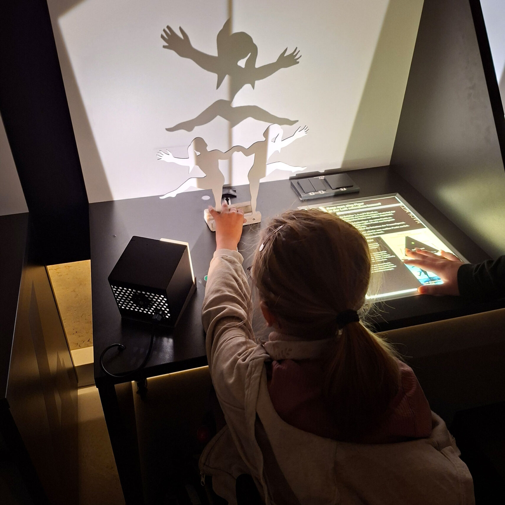 Ein Kind an der Martinus-Schule beschäftigt sich während der Projekttage mit einer naturwissenschaftlichen Ausstellung auf einem Tisch und verschiebt weiße Pappfiguren, um kreative Schattenbilder an eine Wand zu werfen, die von einem kleinen, würfelförmigen Projektor beleuchtet wird. Eine andere Hand berührt eine Informationstafel in der Nähe. Ein Kind an der Martinus-Schule beschäftigt sich während der Projekttage mit einer naturwissenschaftlichen Ausstellung auf einem Tisch und verschiebt weiße Pappfiguren, um kreative Schattenbilder an eine Wand zu werfen, die von einem kleinen, würfelförmigen Projektor beleuchtet wird. Eine andere Hand berührt eine Informationstafel in der Nähe.